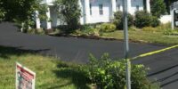 Freshly seal-coated driveway at a white residential home with Pinwheel Property Maintenance LLC sign displayed, showcasing high-quality asphalt work.