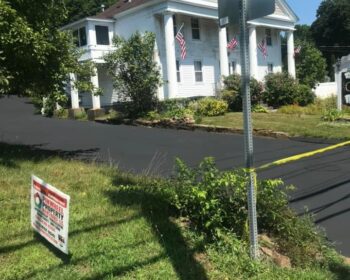 Freshly seal-coated driveway at a white residential home with Pinwheel Property Maintenance LLC sign displayed, showcasing high-quality asphalt work.
